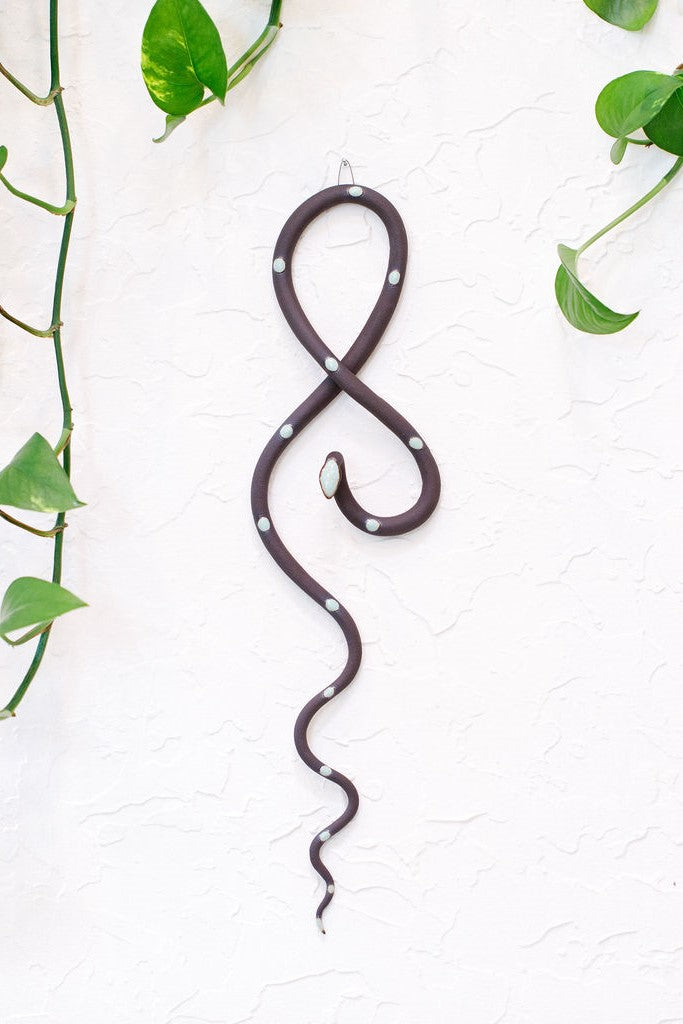 Dark chocolate speckled clay vertical wall snake sculpture with light turquoise glazed head and dots. This colorway is Stevie. The top of the snake crosses like a figure eight and the tail hangs down in an organic s curve. It hangs vertically from a small metal loop on a white wall with vining pothos.