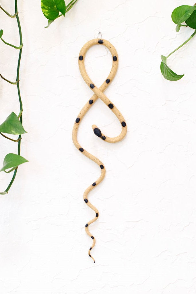 Tan speckled clay vertical wall snake sculpture with black glazed head and dots. This colorway is Sammy. The top of the snake crosses like a figure 8 and the tail hangs down in an organic s curve. It hangs vertically from a small metal loop on a white wall with vining pothos.