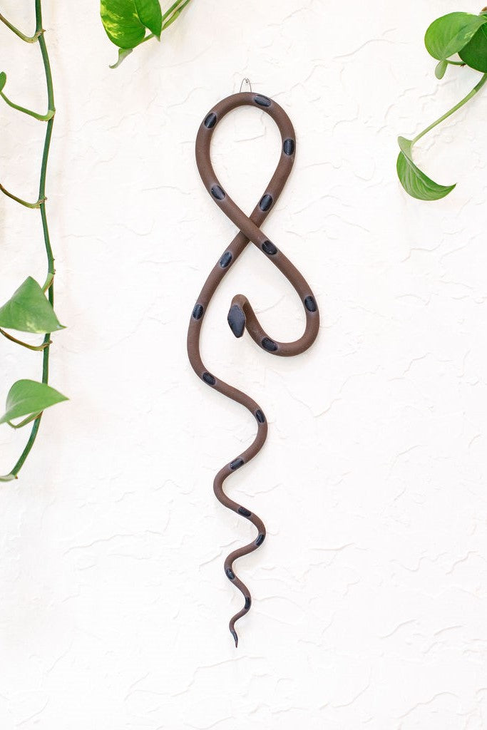 Dark brown clay vertical wall snake sculpture with black glazed head and dots. This colorway is Saul. The top of the snake crosses like a figure 8 and the tail hangs down in an organic s curve. It hangs vertically from a small metal loop on a white wall with vining pothos.
