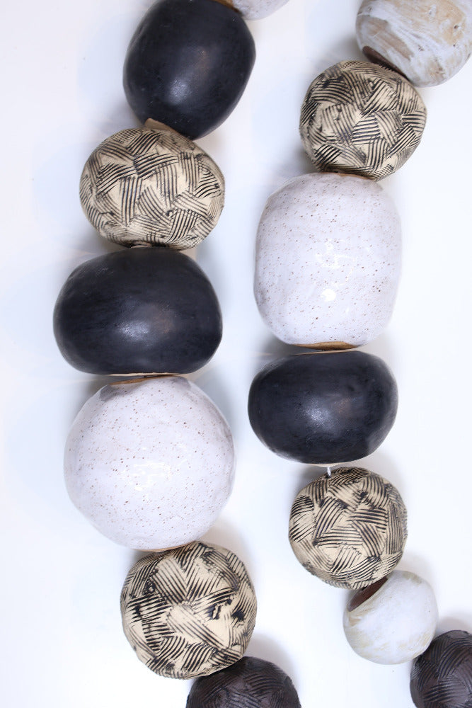 Two strands of ceramic balls laying parallel to each other in a slight curve on a white surface. The ceramic balls are glazed in varying shades of black, white and tan with different textures etched into some of the balls. The balls are irregularly shaped also vary in size.
