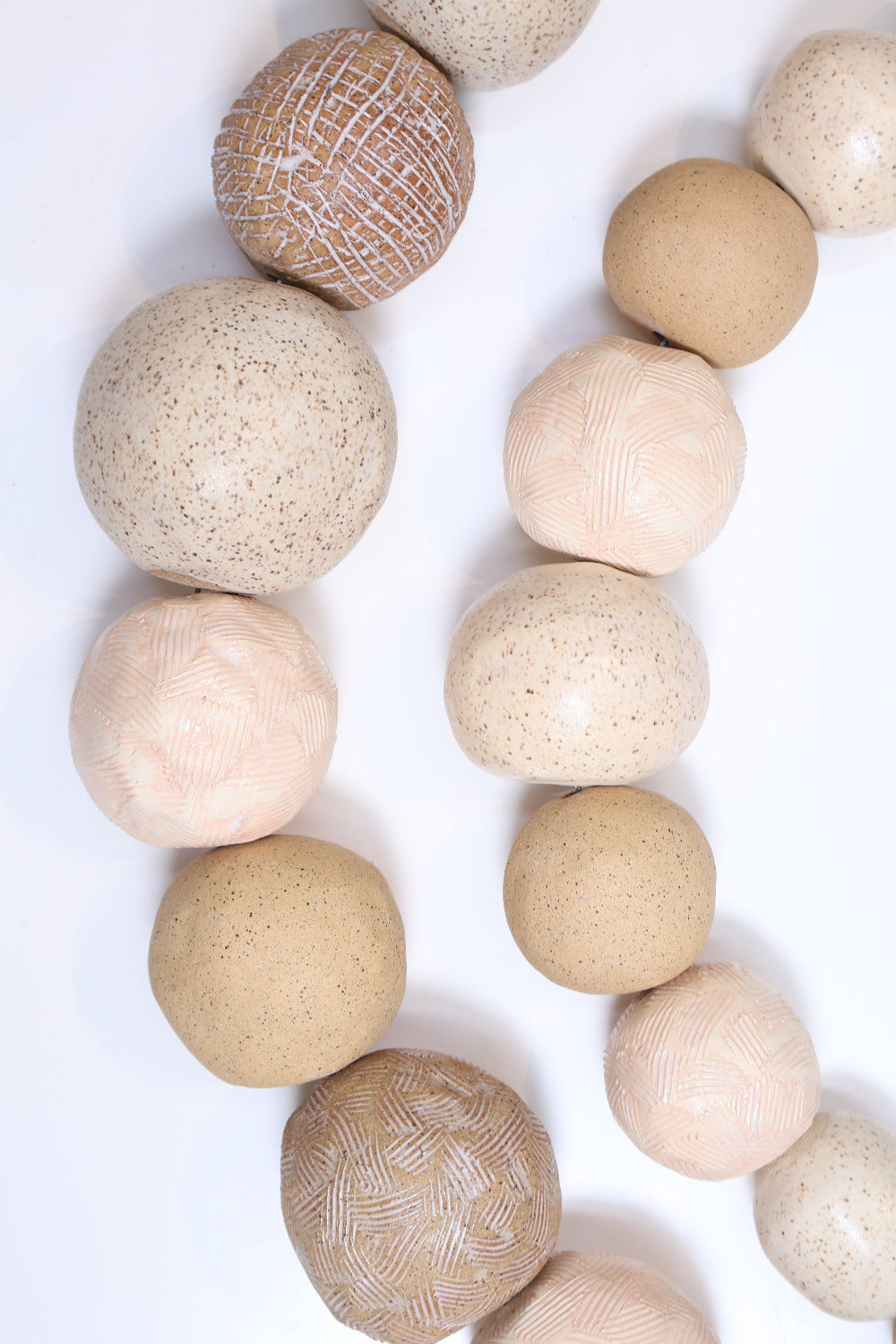 Two strands of ceramic balls laying parallel to each other in a slight curve on a white surface. The ceramic balls are glazed in varying shades of tan with different textures etched into some of the balls. The balls also vary in size.