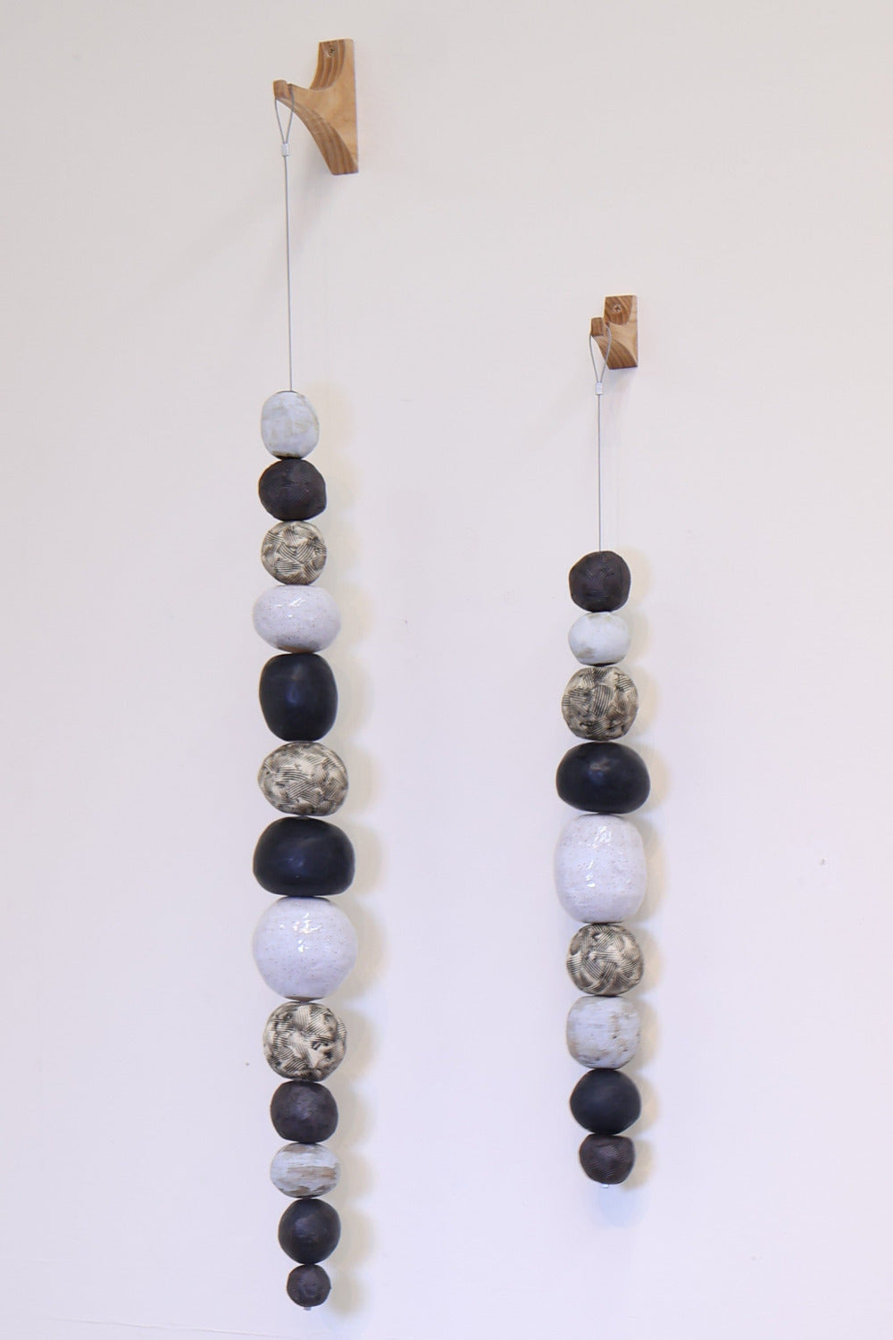 Two decorative strands of irregularly shaped ceramic balls of varying sizes hanging from wooden wall hooks. The color scheme of these strands is speckled white, matte black and t