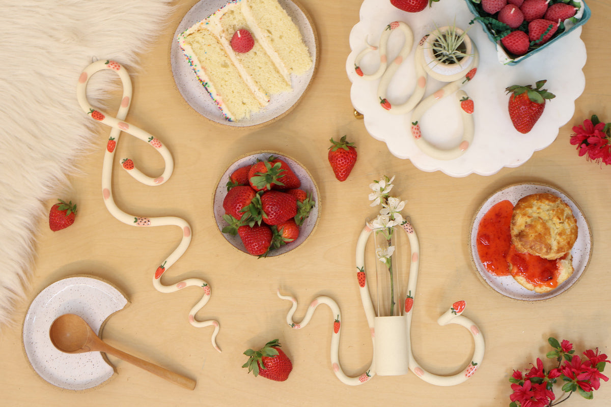 A styled flat lay of strawberry ceramic snakes. The snakes are ivory clay with glazed strawberries and pink dots. They are surrounded by fresh strawberries, a slice of cake and a biscuit with strawberry jam.