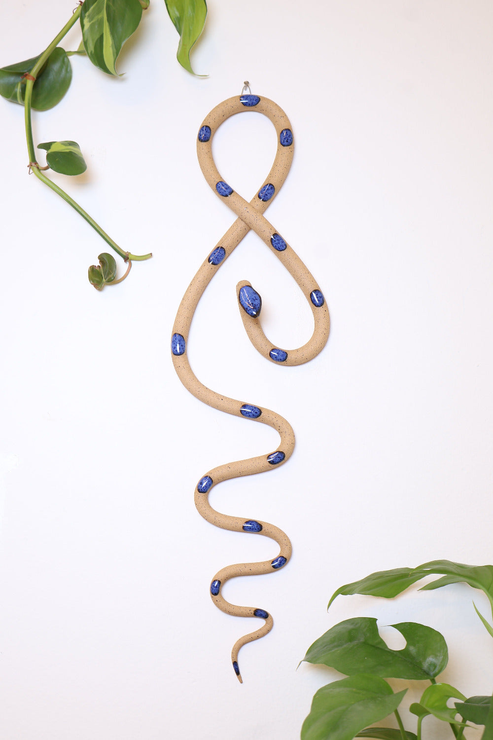 Tan speckled clay vertical wall snake sculpture with dark blue glazed head and dots. This colorway is Selena. The top of the snake crosses like a figure 8 and the tail hangs down in an organic s curve. It hangs vertically from a small metal loop on a white wall with vining pothos.