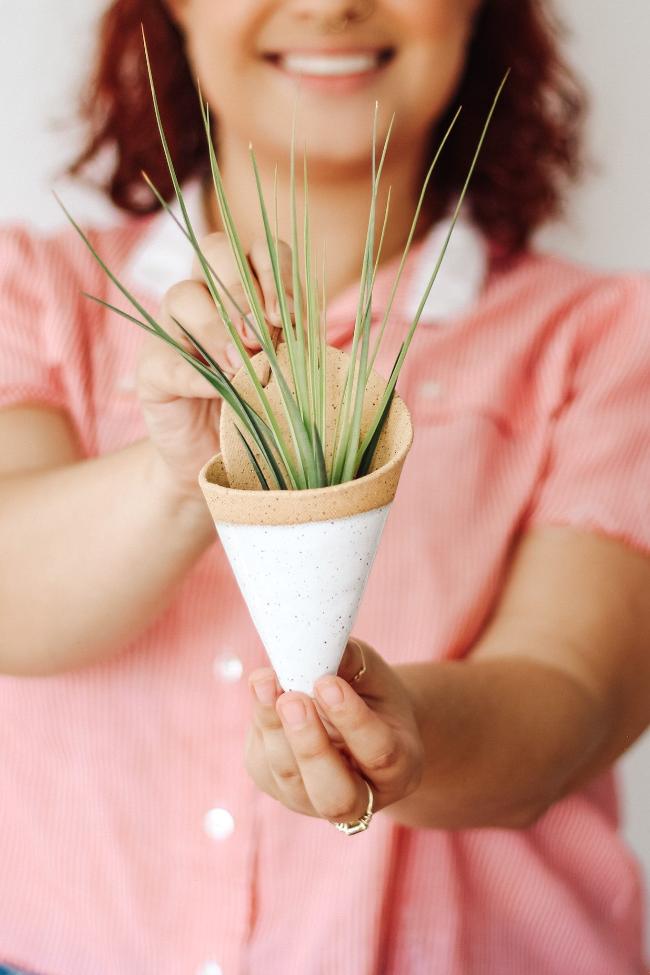 Speckled clay ceramic cone-shaped planter with dipped white glaze styled with a spikey green air plant. The cone is being held by a young smiling woman showing the faux leather loop at the top for hanging the cone.
