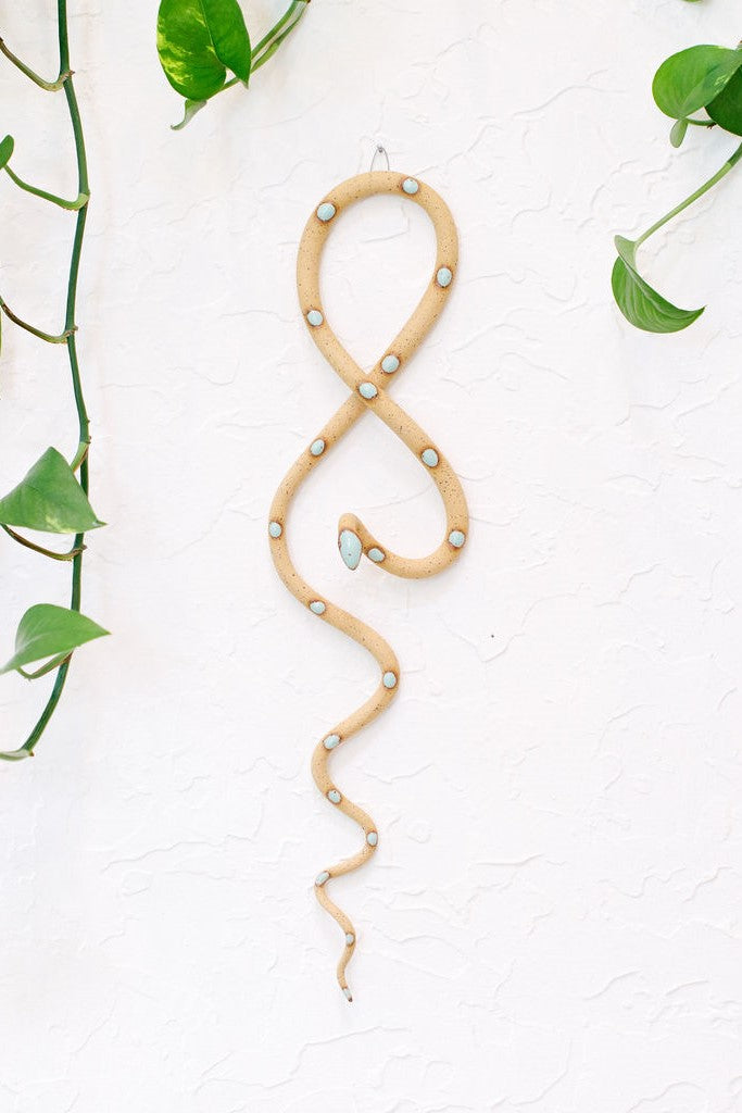 Tan speckled clay vertical wall snake sculpture with light turquoise glazed head and dots. This colorway is Selma. The top of the snake crosses like a figure 8 and the tail hangs down in an organic s curve. It  hangs vertically from a small metal loop on a white wall with vining pothos. 