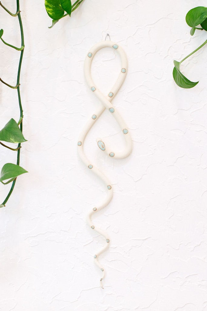 Ivory clay vertical wall snake sculpture with light turquoise glazed head and dots. This colorway is Sylvia. The top of the snake crosses like a figure 8 and the tail hangs down in an organic s curve. It hangs vertically from a small metal loop on a white wall with vining pothos. 