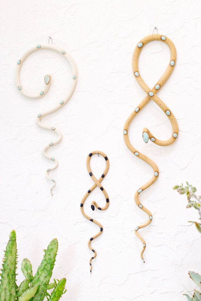 Forest curated set of three wall ceramic snakes. From left to right hanging on a wall a medium Saul wall snake (black clay and black glaze color way), a Sammy mini wall snake (speckled tan and black glaze colorway) and a Stevie wall snake (black clay and turquoise glaze colorway). 
