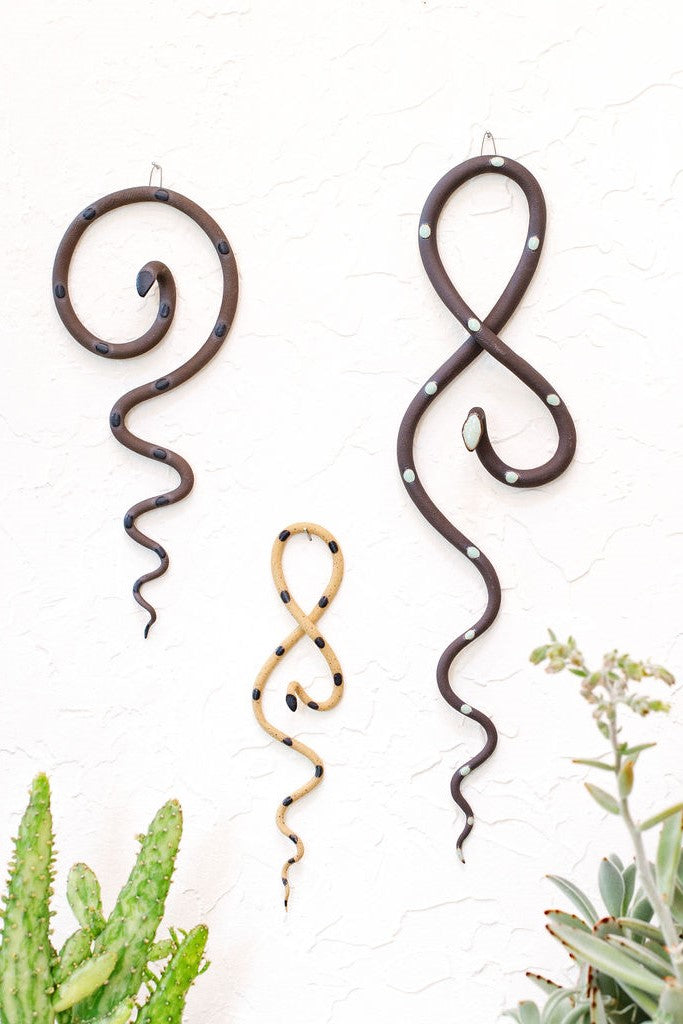 Forest curated set of three wall ceramic snakes. From left to right hanging on a wall a medium Saul wall snake (black clay and black glaze color way), a Sammy mini wall snake (speckled tan and black glaze colorway) and a Stevie wall snake (black clay and turquoise glaze colorway). 