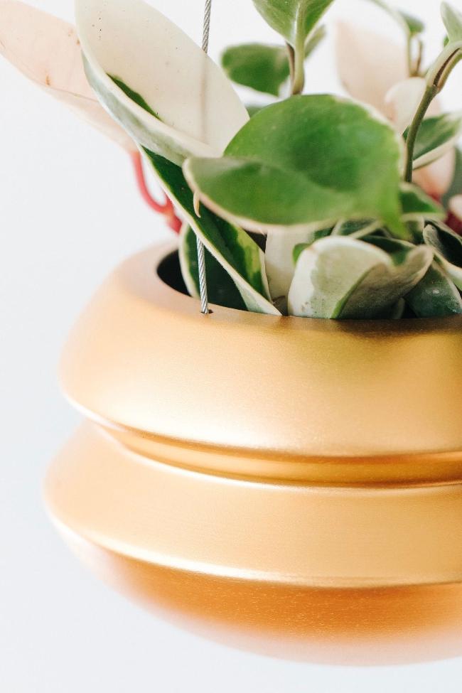 Close up of the double fold accordion like design of the large brass hanging halo planter. The halo is holding a vining white, pink and green plant. 