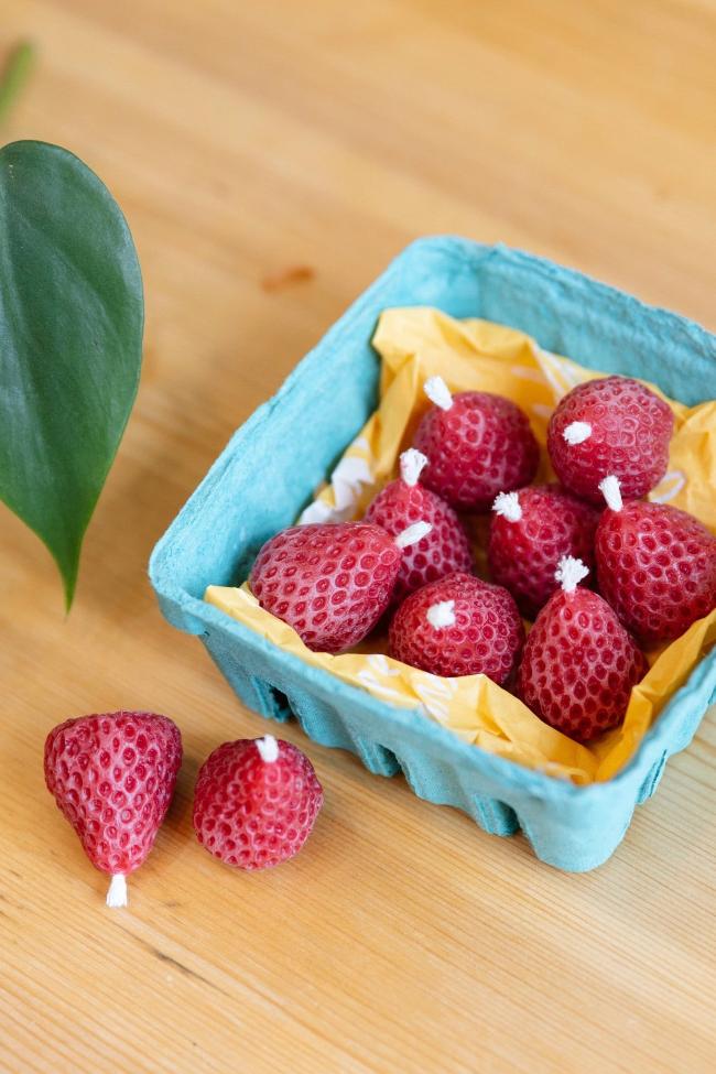 Eight red strawberry shaped beeswax candles nestled in a teal berry box with two candles outside of the box. 