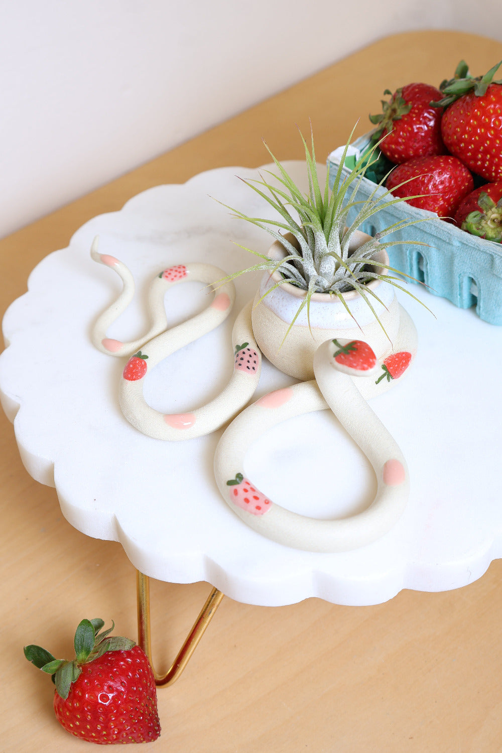 Decorative snake-shaped table sculpture with a small attached pot. The snake has a strawberry and pink dot pattern and sits on a white stand surrounded by fresh strawberries. The pot holds a small air plant.