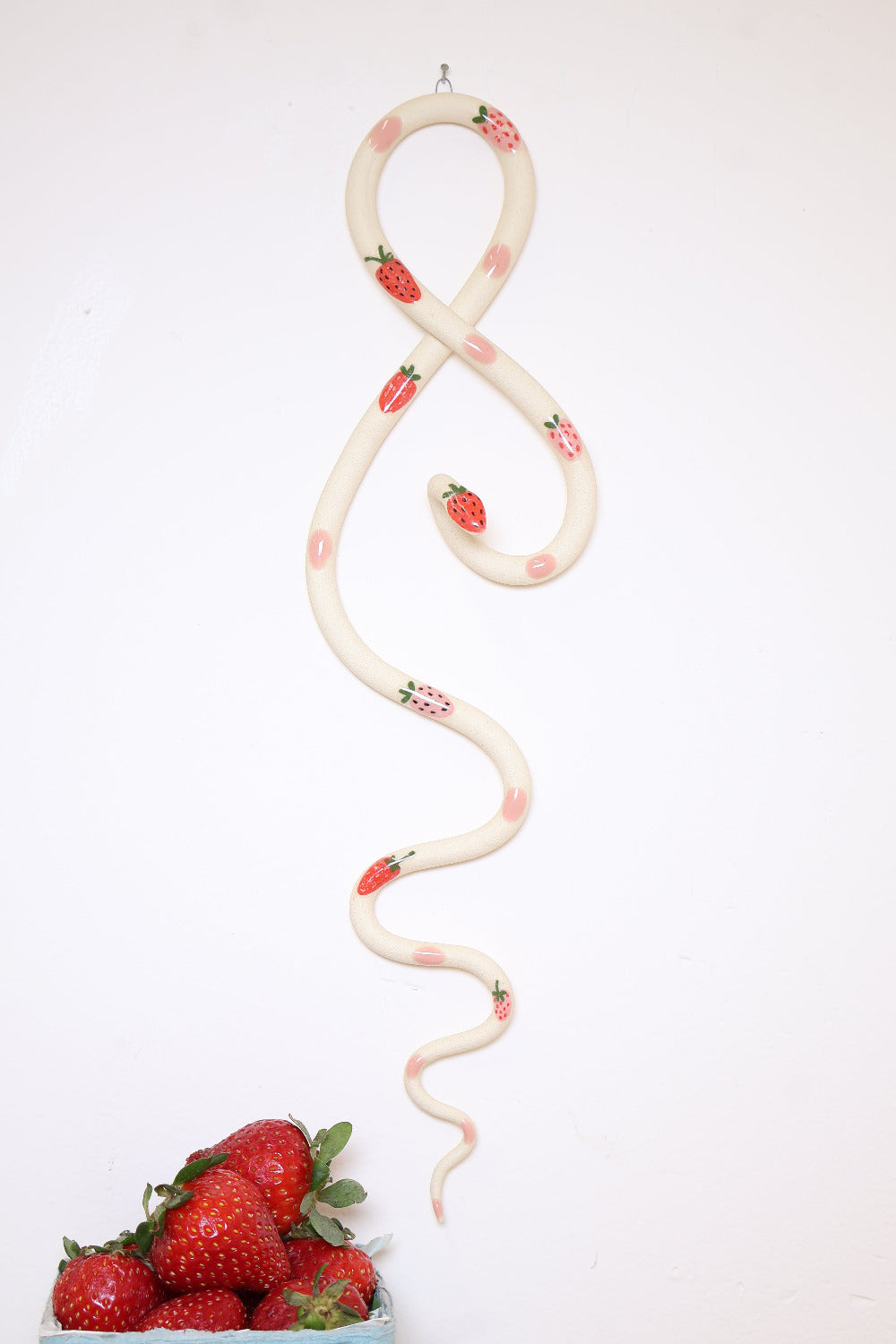 Decorative snake with strawberry and pink dot pattern on a white wall next to a bowl of fresh strawberries.