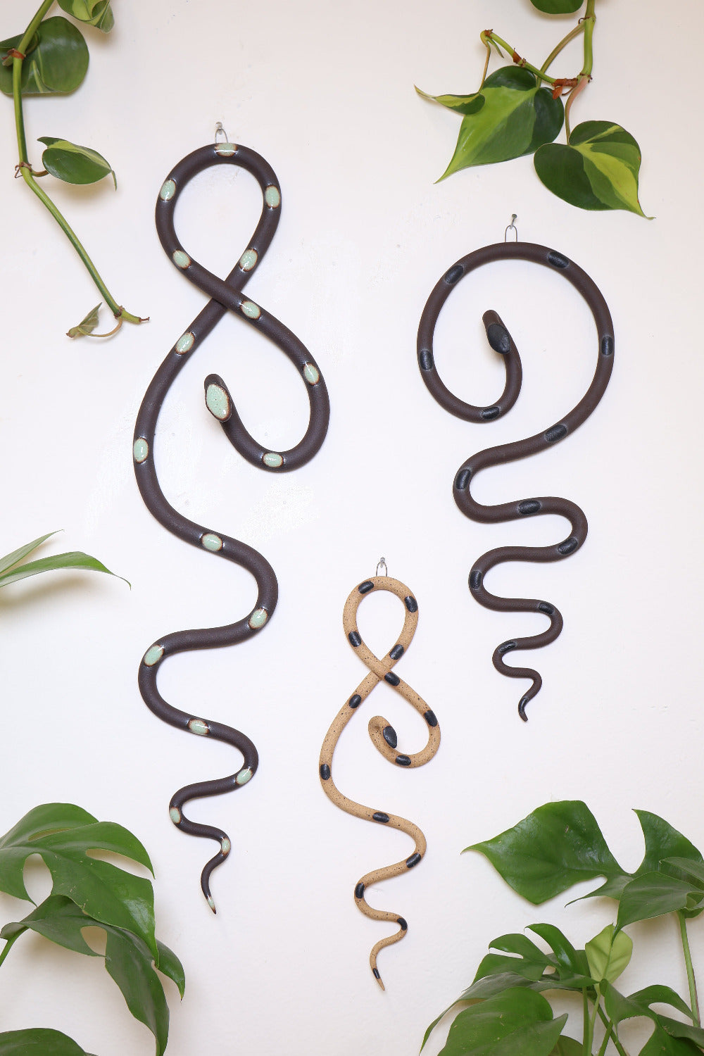 Forest curated set of three wall ceramic snakes. From left to right hanging on a wall is a Stevie wall snake (black clay and turquoise glaze colorway), a mini Sammy wall snake (speckled tan and black glaze colorway), and a medium Saul wall snake (black clay and black glaze color way).