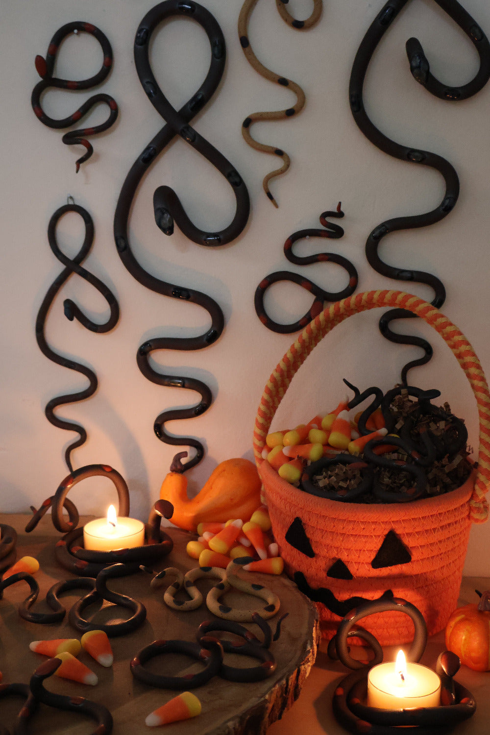 Collection of the fall limited edition snakes in dim lighting, hung on the wall and spread on the table top with candy corn and a woven jack-o-lantern basket. The coiled snakes are holding lit tealights to create a candlelight mood. 