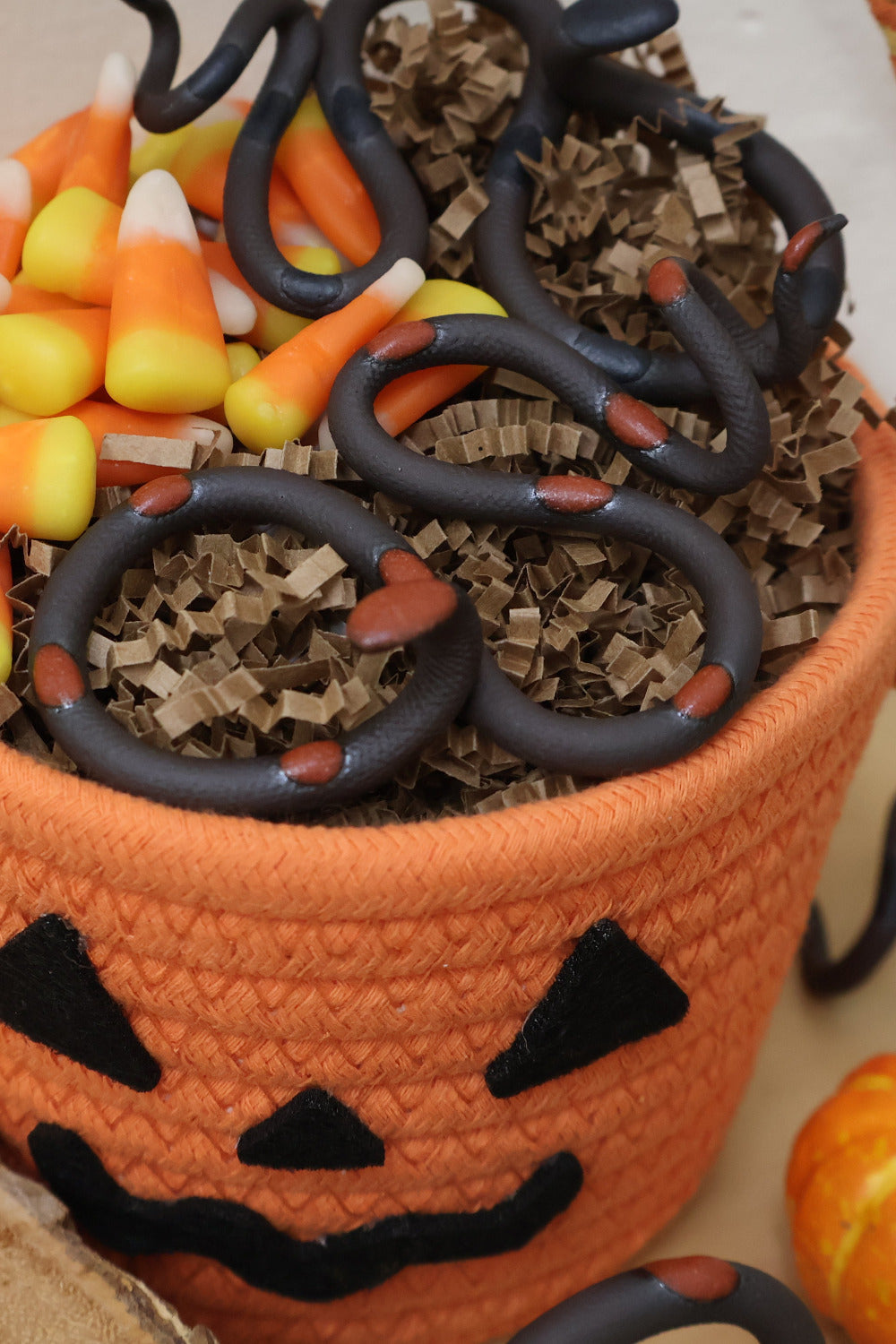 An orange woven Halloween basket with a jack-o'-lantern face, filled with candy corn and a Squash and Saul ceramic small snake.