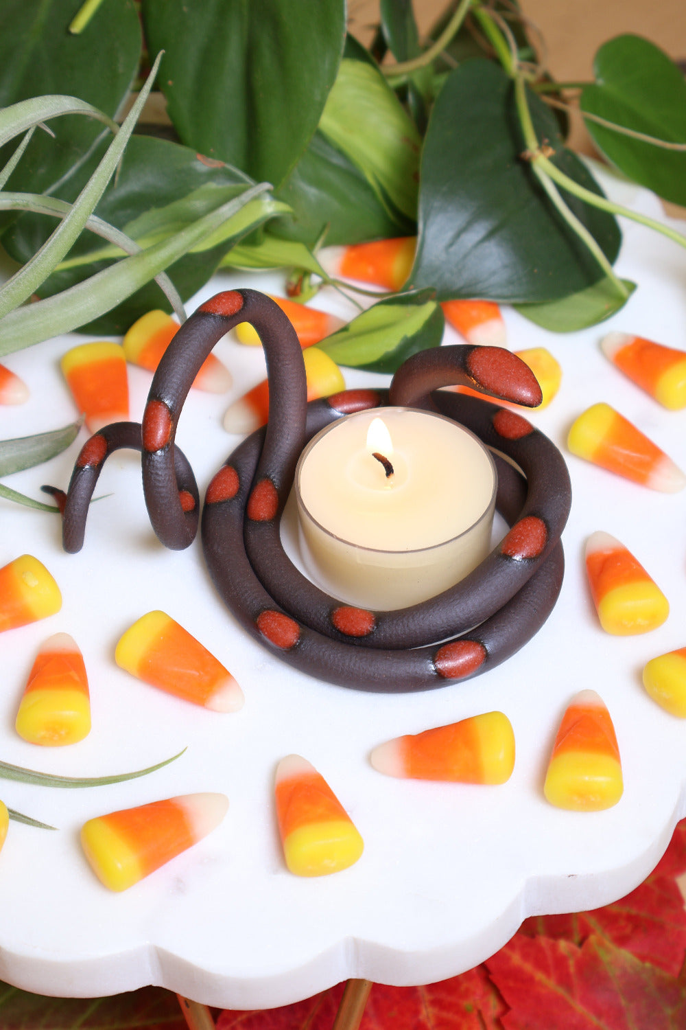 Organically coiled black clay snake with red-orange glazed head and dots. This colorway is Squash. Styled with a beeswax tea light that sits in the center of the coil. The tealight is lit and the snake is surrounded by festive candy corn.