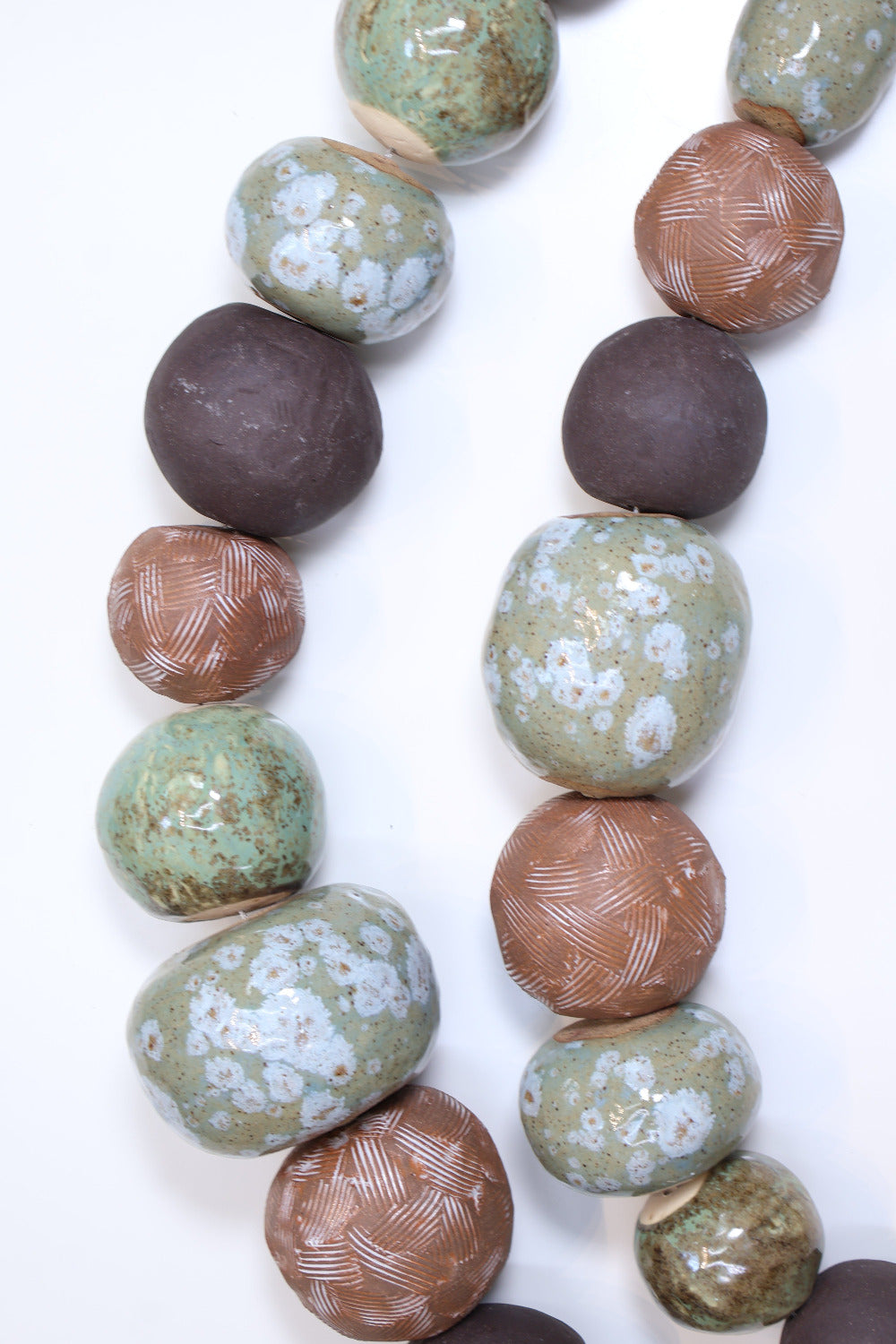 Two strands of irregular ceramic balls laying parallel to each other in a slight curve on a white surface. The ceramic balls are glazed in varying shades of earthy green, brown, and black with different  textures etched into some of the balls. The balls also vary in size. 