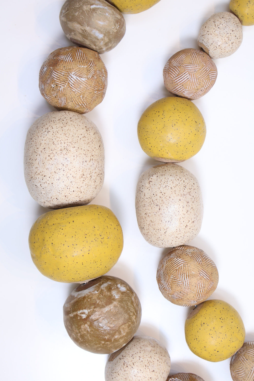 Two strands of ceramic balls laying parallel to each other in a slight curve on a white surface. The ceramic balls are glazed in varying shades of tan and yellow with different  textures etched into some of the balls. The balls also vary in size. 
