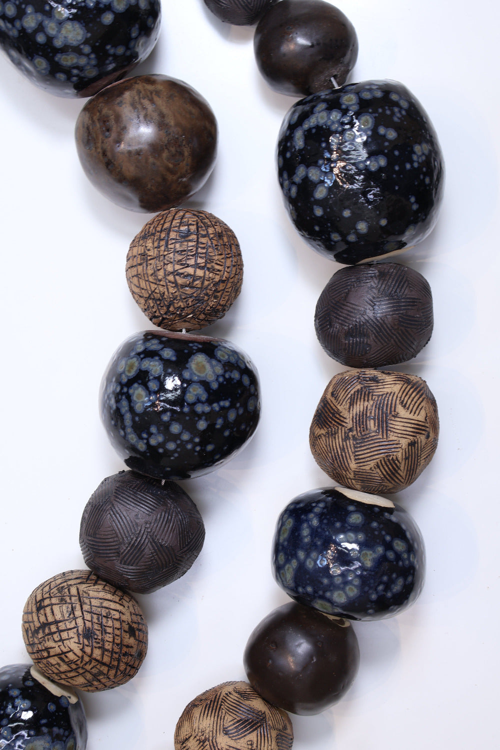 Two strands of ceramic balls laying parallel to each other in a slight curve on a white surface. The ceramic balls are glazed in varying shades of black and brown with different  textures etched into some of the balls. The balls also vary in size. 
