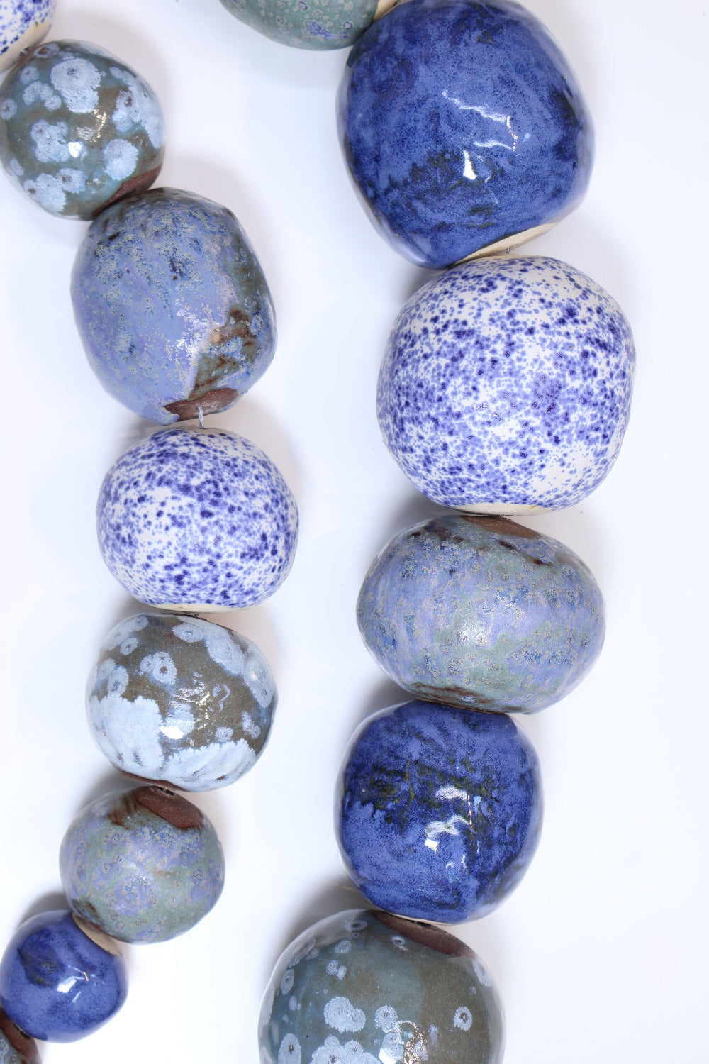 Two strands of ceramic balls laying parallel to each other in a slight curve on a white surface. The ceramic balls are glazed in varying shades of blues and greens with different  textures etched into some of the balls. The balls also vary in size. 