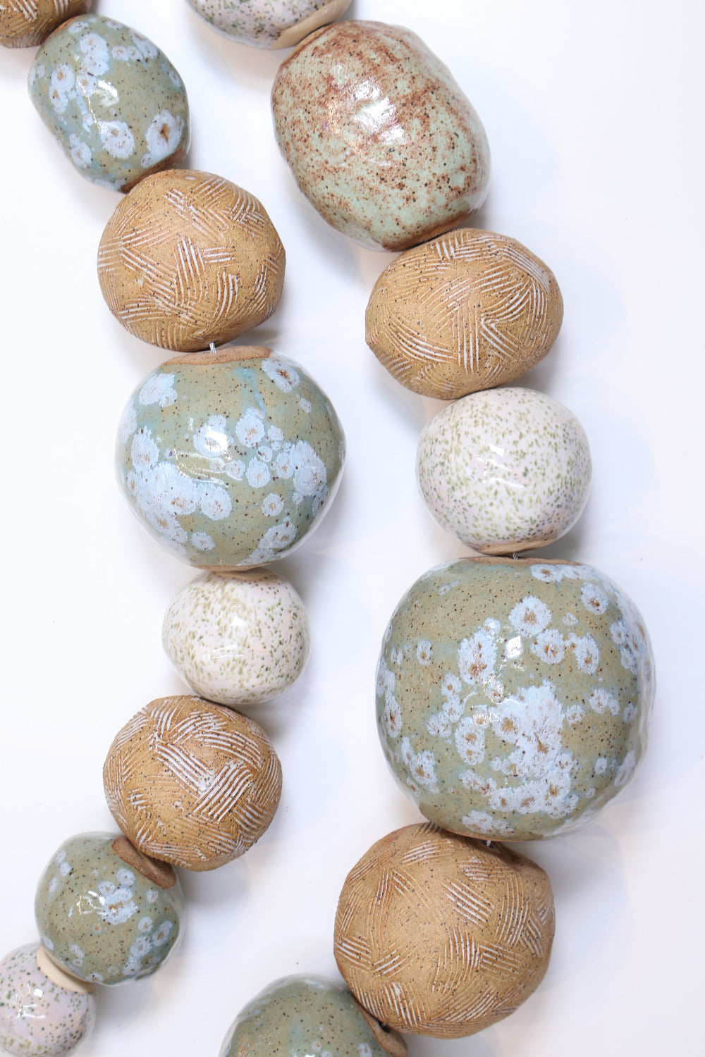 Two strands of ceramic balls laying parallel to each other in a slight curve on a white surface. The ceramic balls are glazed in varying shades of tan, white and light green with different textures etched into some of the balls. The balls also vary in size. 