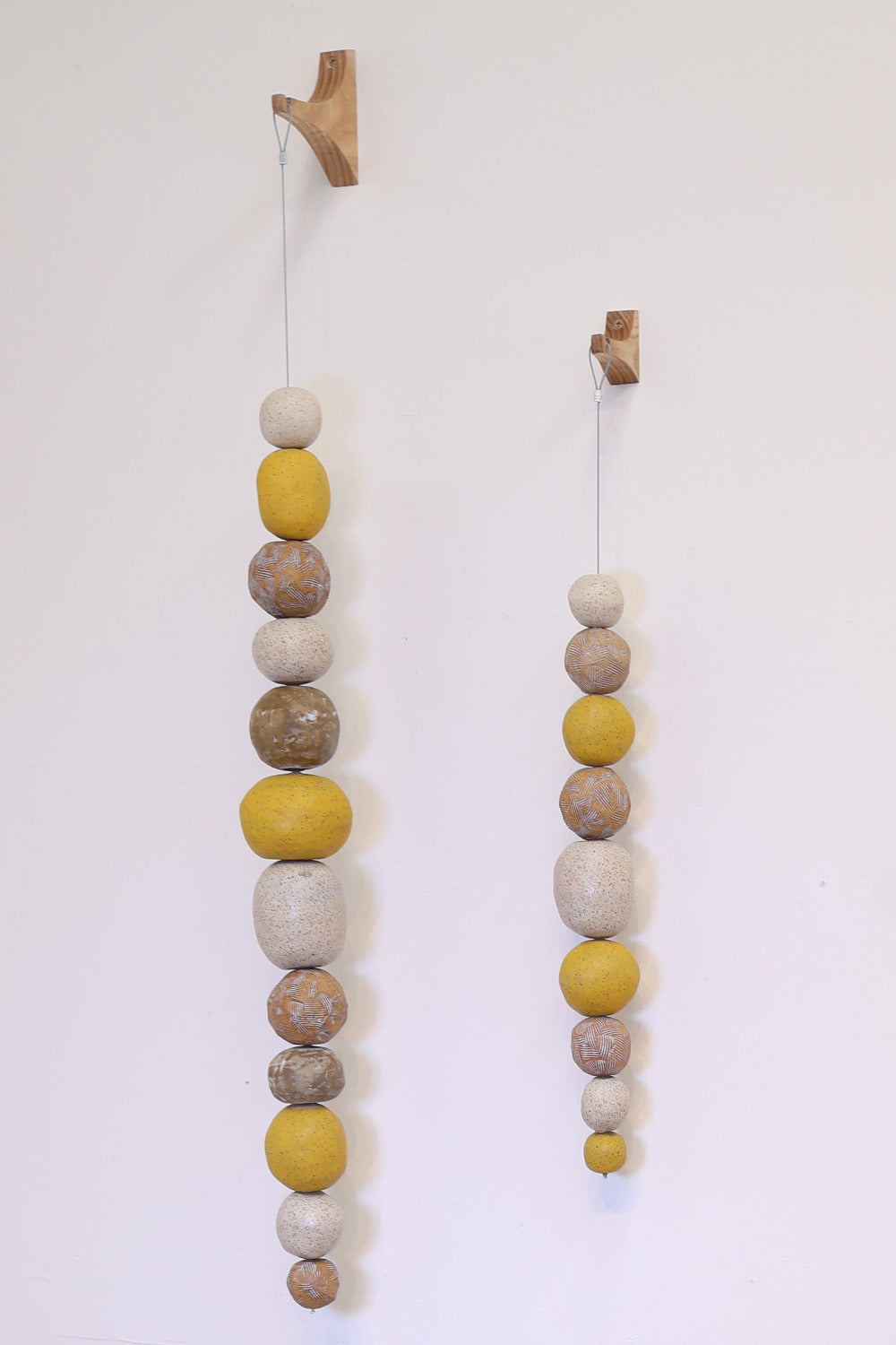 Two decorative strands of irregularly shaped ceramic balls of varying sizes strung along a steel cable, hanging from wooden wall hooks. The color scheme of these strands is speckled white, browns and yellow.