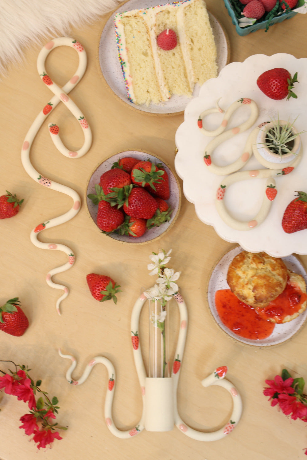 Flat lay of the strawberry ceramic snake collection with three difference snake shapes. The snakes are surrounded by strawberries a slice of cake, red flowers and a jam covered biscuit. 