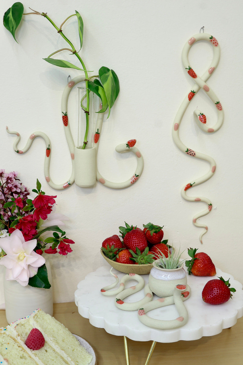 Flat lay of the strawberry ceramic snake collection with three difference snake shapes. The snakes are surrounded by strawberries a slice of cake, red flowers and a jam covered biscuit. 