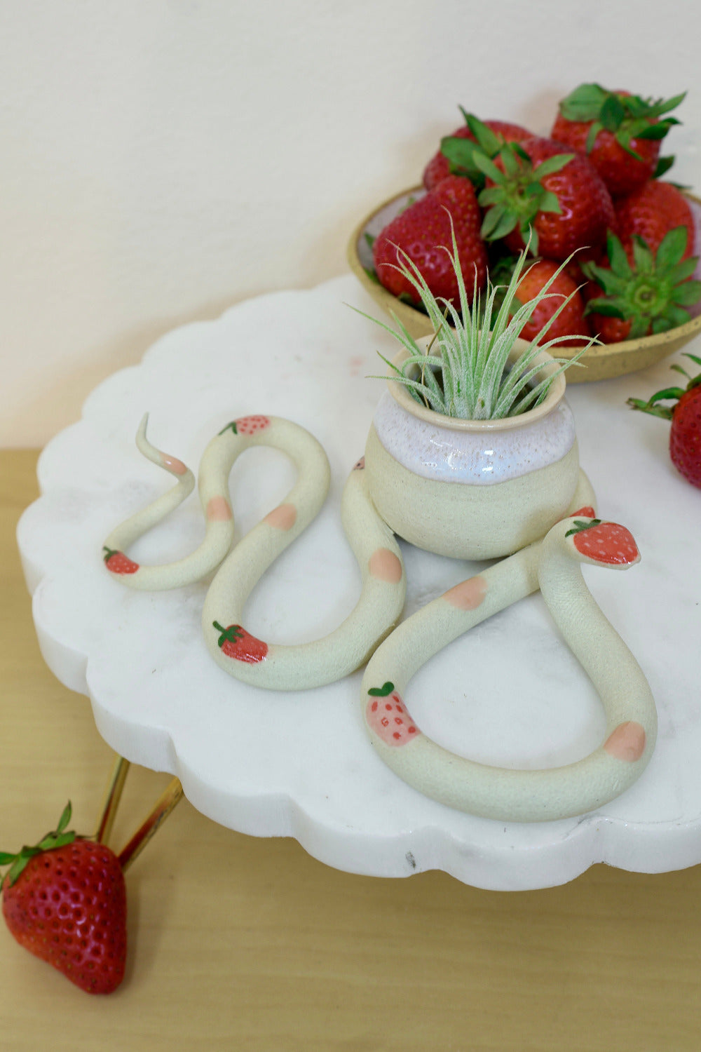 A whimsical ceramic snake with a little decorative pot adorned with strawberry accents and pink dots. The snake is styled with an air plant and a bowl of fresh strawberries sits nearby. 