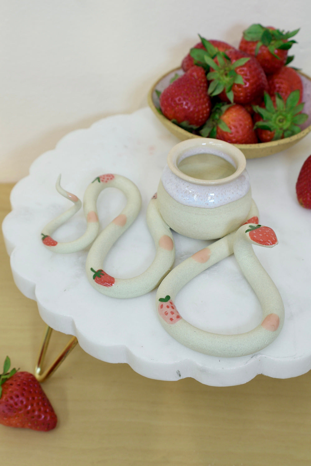Decorative snake-shaped table sculpture with a small attached pot. The snake has a strawberry and pink dot pattern and sits on a white stand surrounded by fresh strawberries.