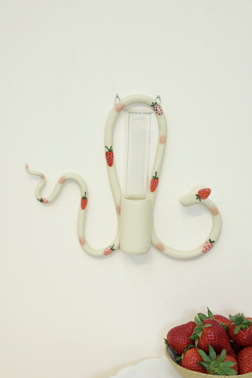 An organic s-shaped white ceramic wall snake with an attached ceramic vase to hold a glass tube for plant propagations. The snake is adorned with red strawberry accents and pink polka dots. 