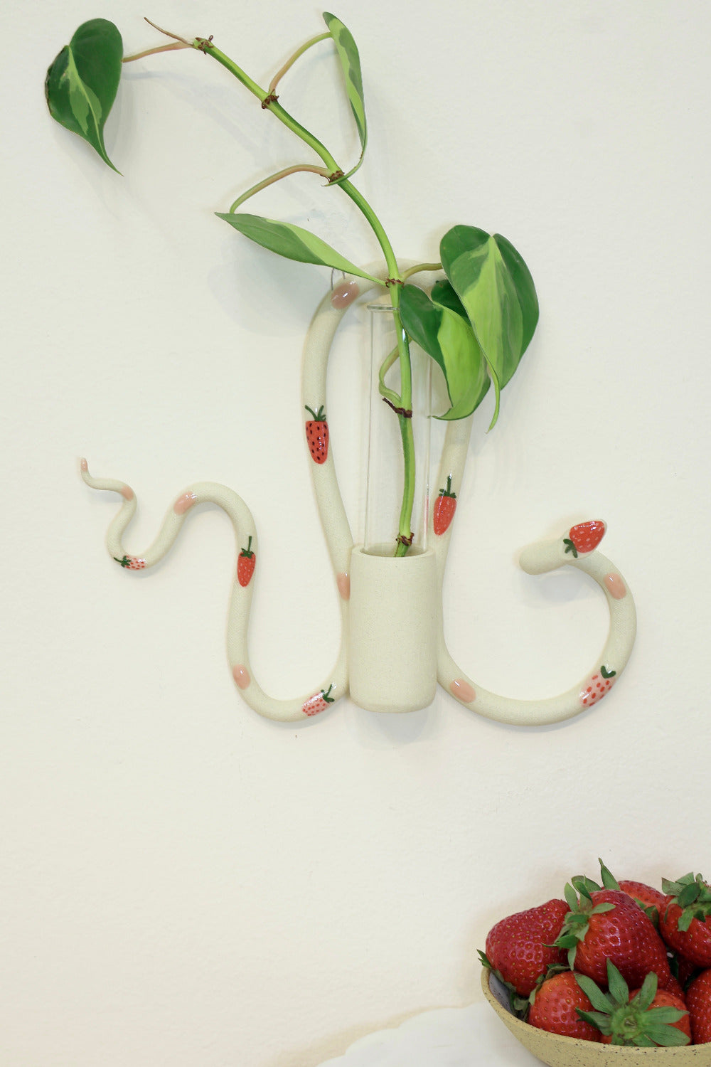 An organic s-shaped white ceramic wall snake with an attached ceramic vase to hold a glass tube for plant propagations. The snake is adorned with red strawberry accents and pink polka dots. The piece holds a green leafy plant, creating a charming, decorative wall piece.