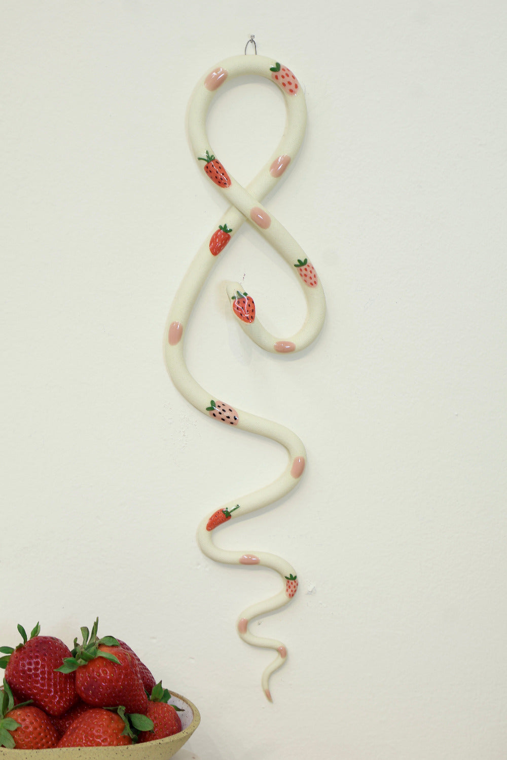 Decorative snake with strawberry and pink dot pattern on a white wall next to a bowl of fresh strawberries.