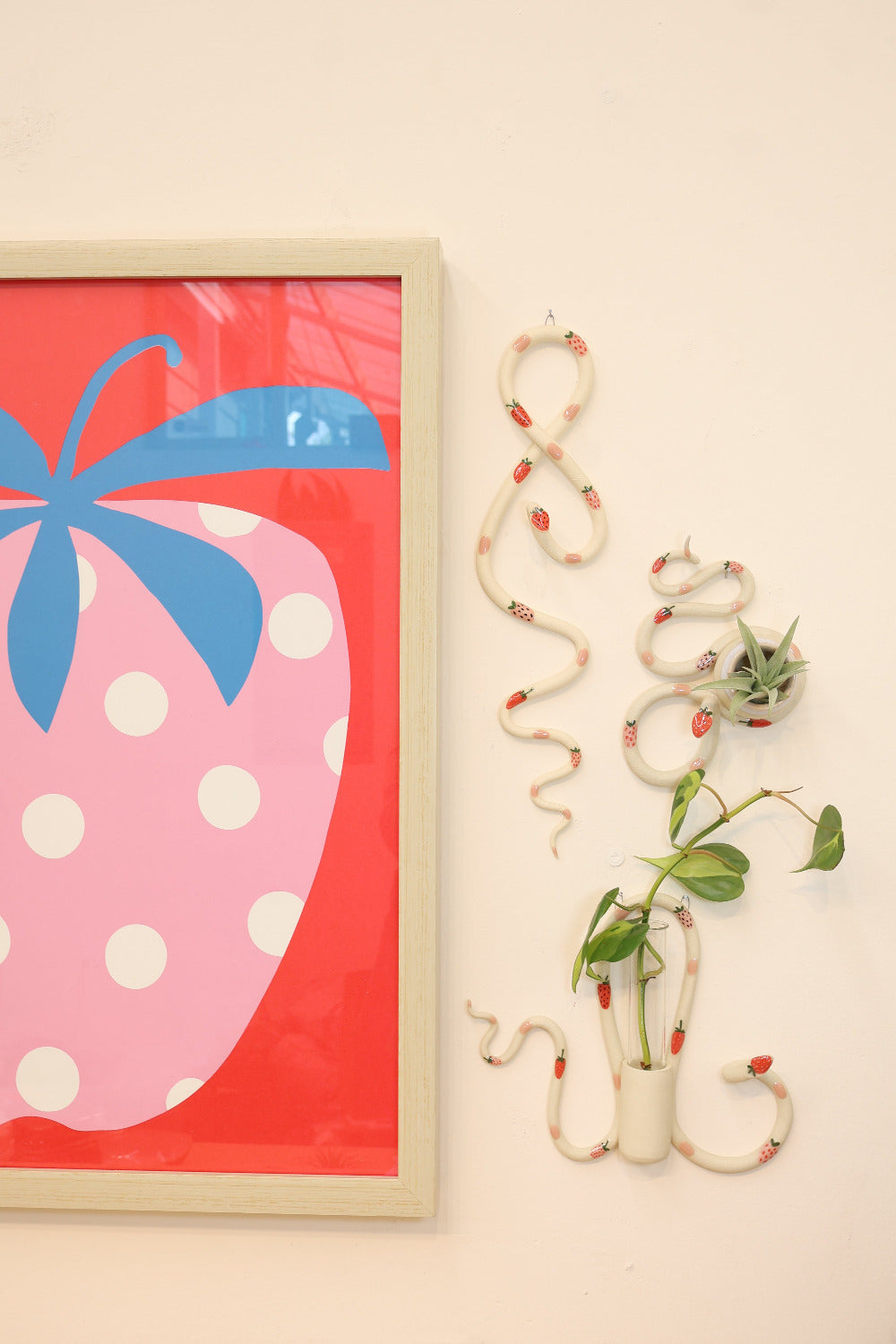 Strawberry wall snake, bud vase and propagation station hanging on a white wall next to a colorful strawberry art print. 