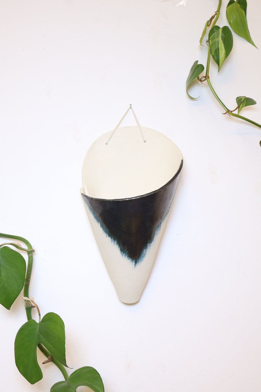 A jumbo ceramic wall planter with a faux leather hanging strap. The planter is shaped like a cone and formed from an ivory clay and has a brushed indigo glaze design. The cone is hanging on a white wall with vining plants.