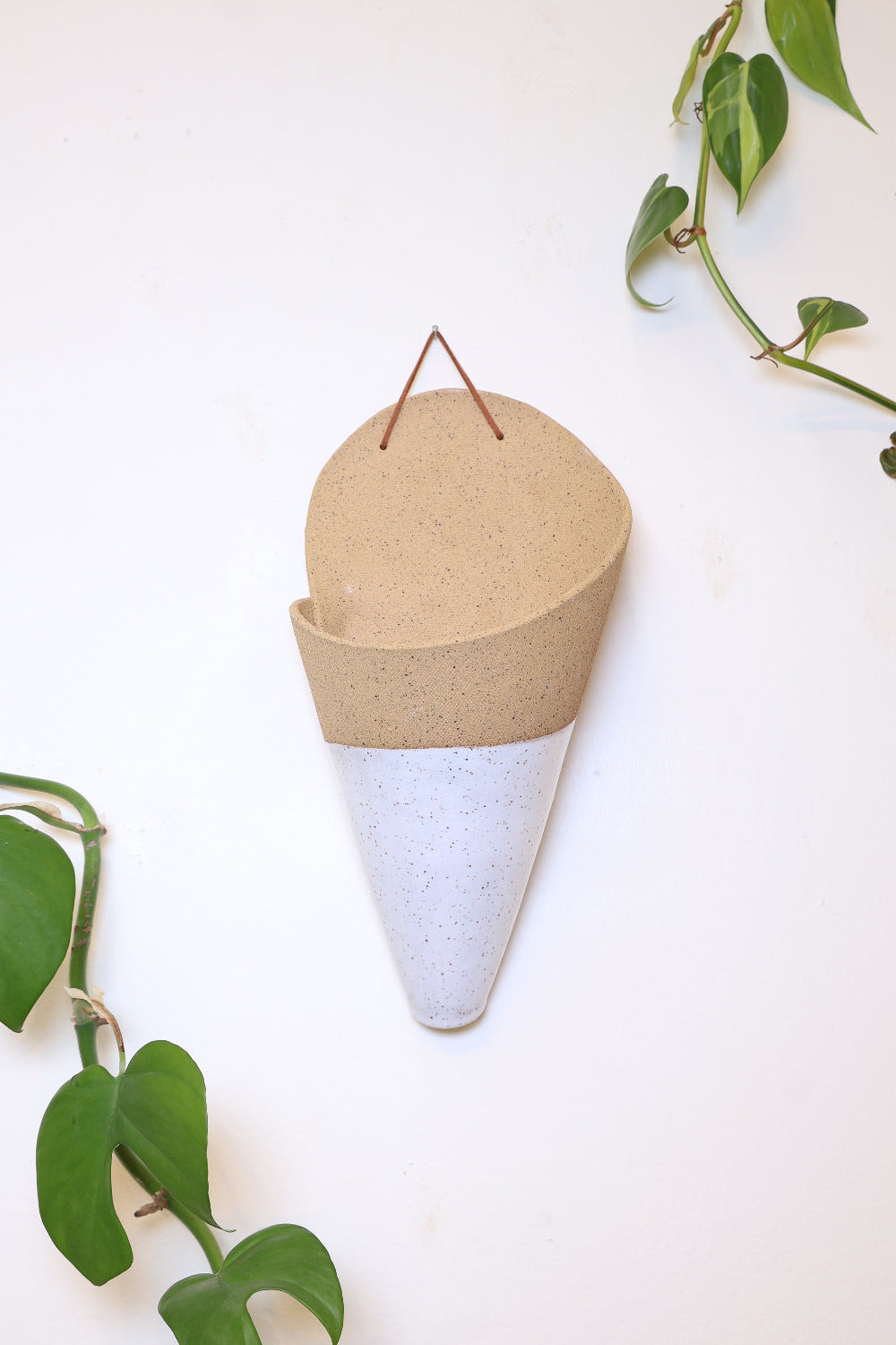 A jumbo ceramic wall planter with a faux leather hanging strap. The planter is shaped like a cone and is made with a tan clay and is dipped in a white speckled glaze. It's styled with a green spikey air plant inside the cone. 
