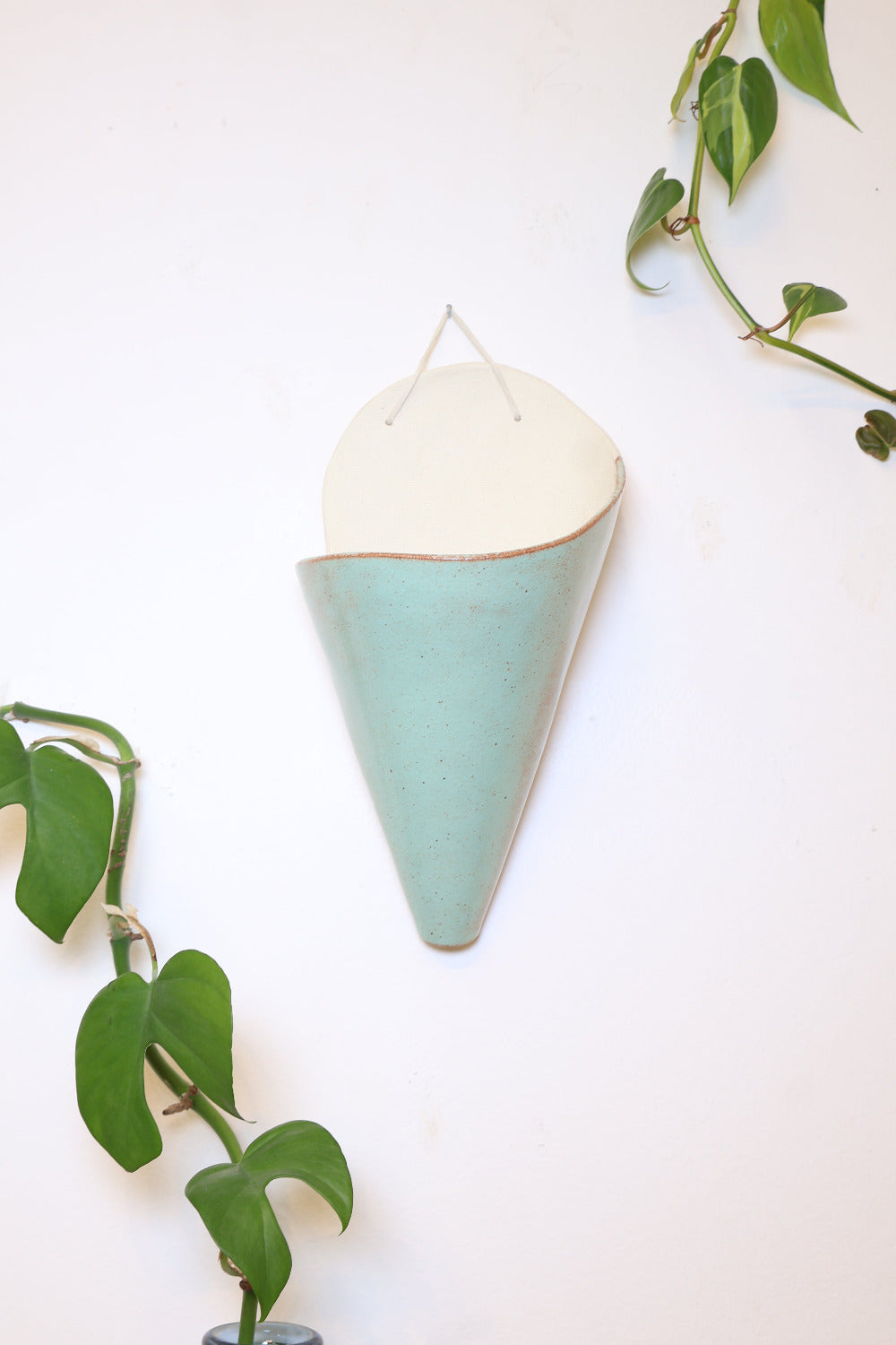 A jumbo ceramic wall planter with a faux leather hanging strap. The planter is shaped like a cone and formed from an ivory clay and is glazed with a light turquoise glaze with warm rust accents. The cone is hanging on a white wall with vining plants.