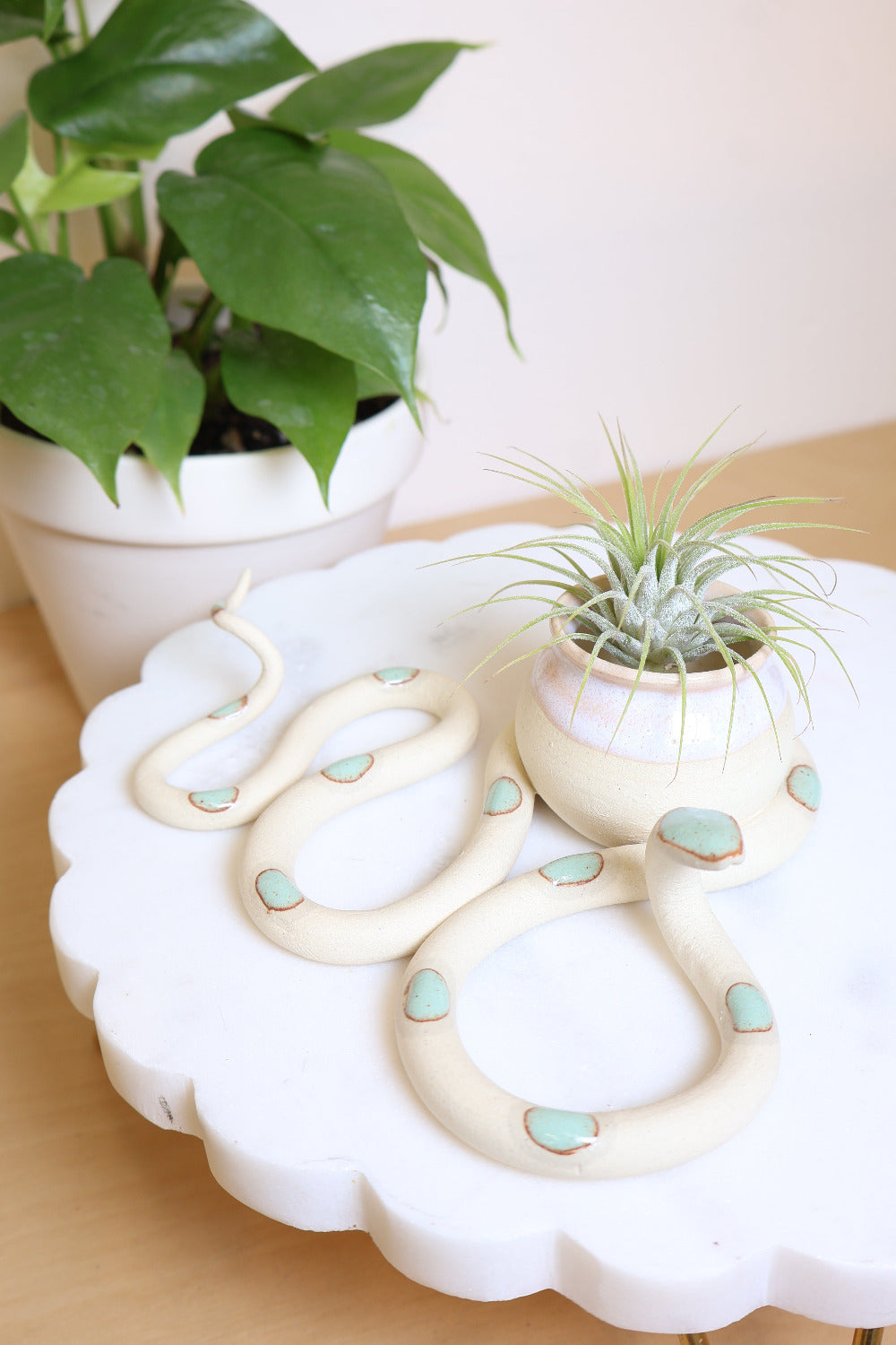 An ivory ceramic snake-shaped sculpture with with green dots and a small bud vase on top. The vase is also ivory with a transparent white glazed top. This colorway is Sylvia. It's styled with an air plant next to a houseplant.
