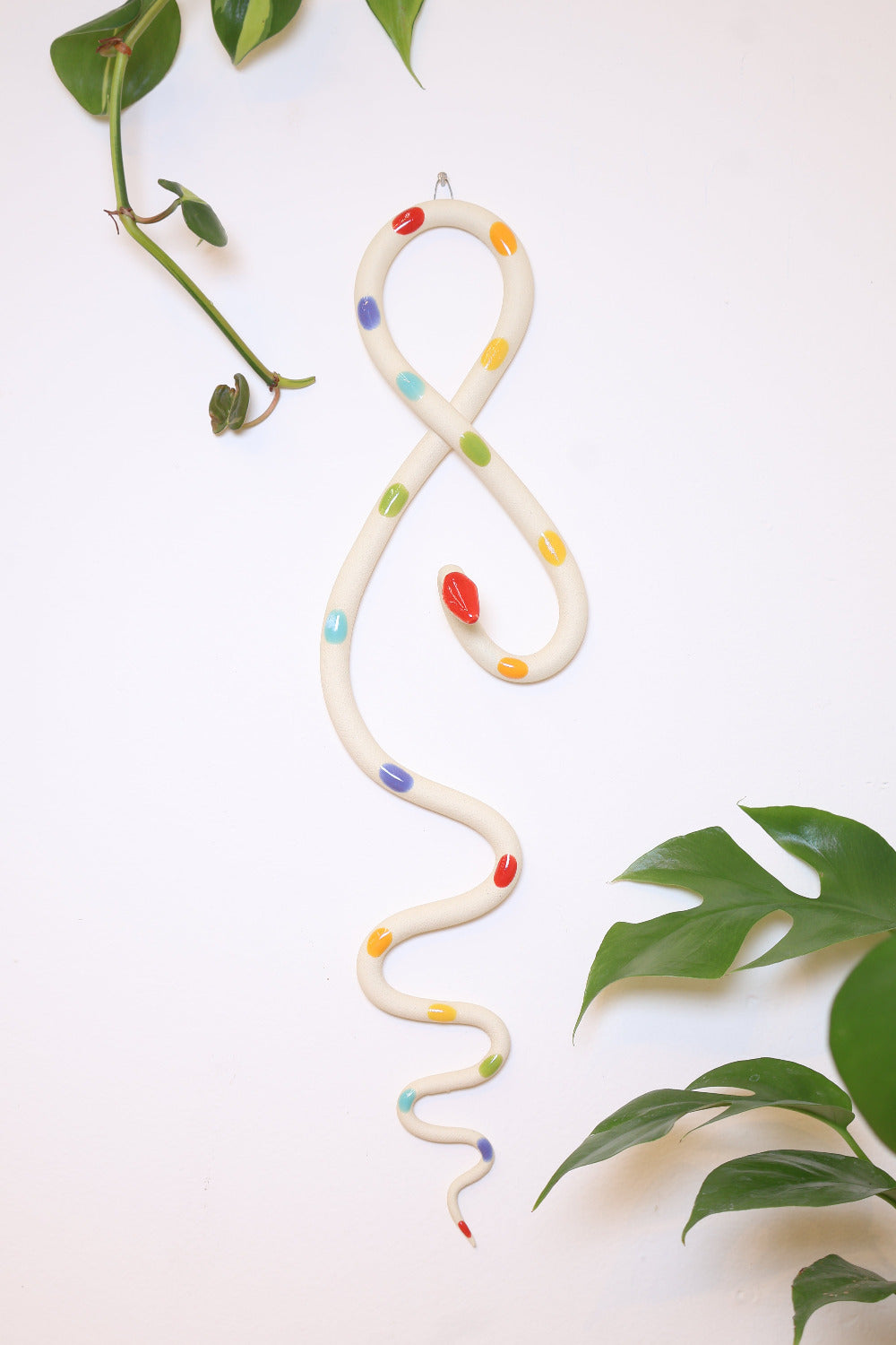 Ivory ceramic wall snake with rainbow glaze dots handing on a white wall with houseplant leaves. 