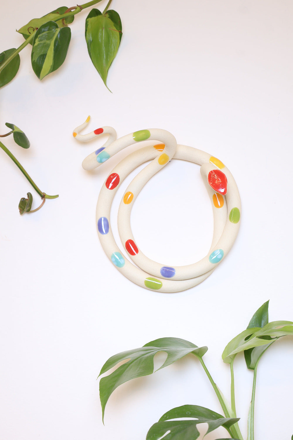 Large ceramic rainbow dotted snake hanging on a white wall with leafy houseplants. 