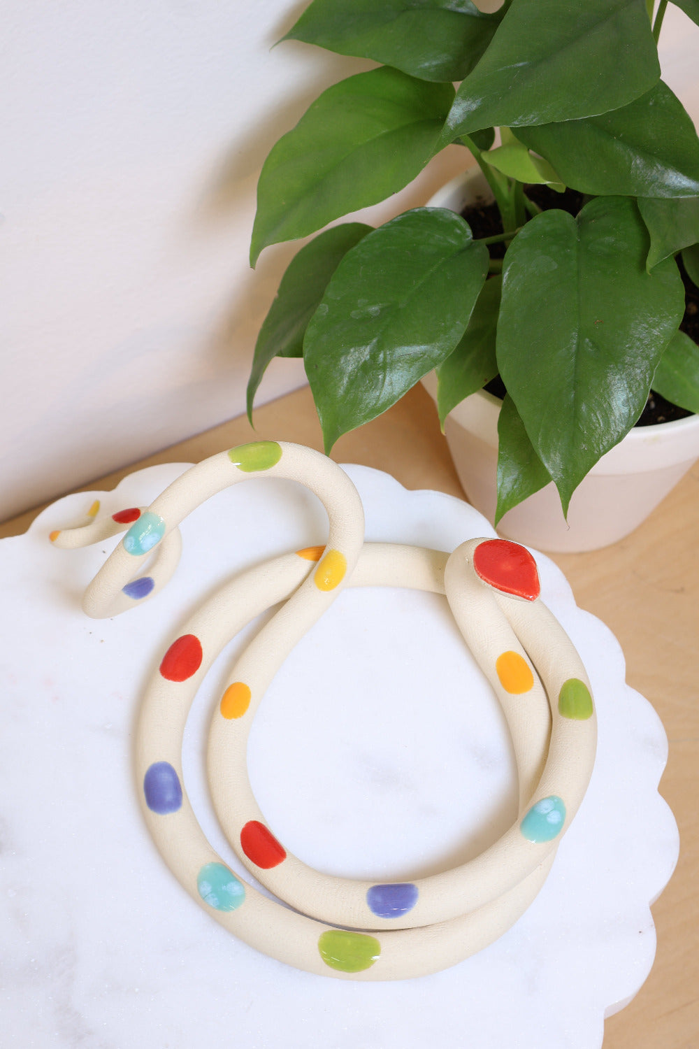 Rainbow dotted ceramic large snake sitting on a marble dish next to a houseplant. 