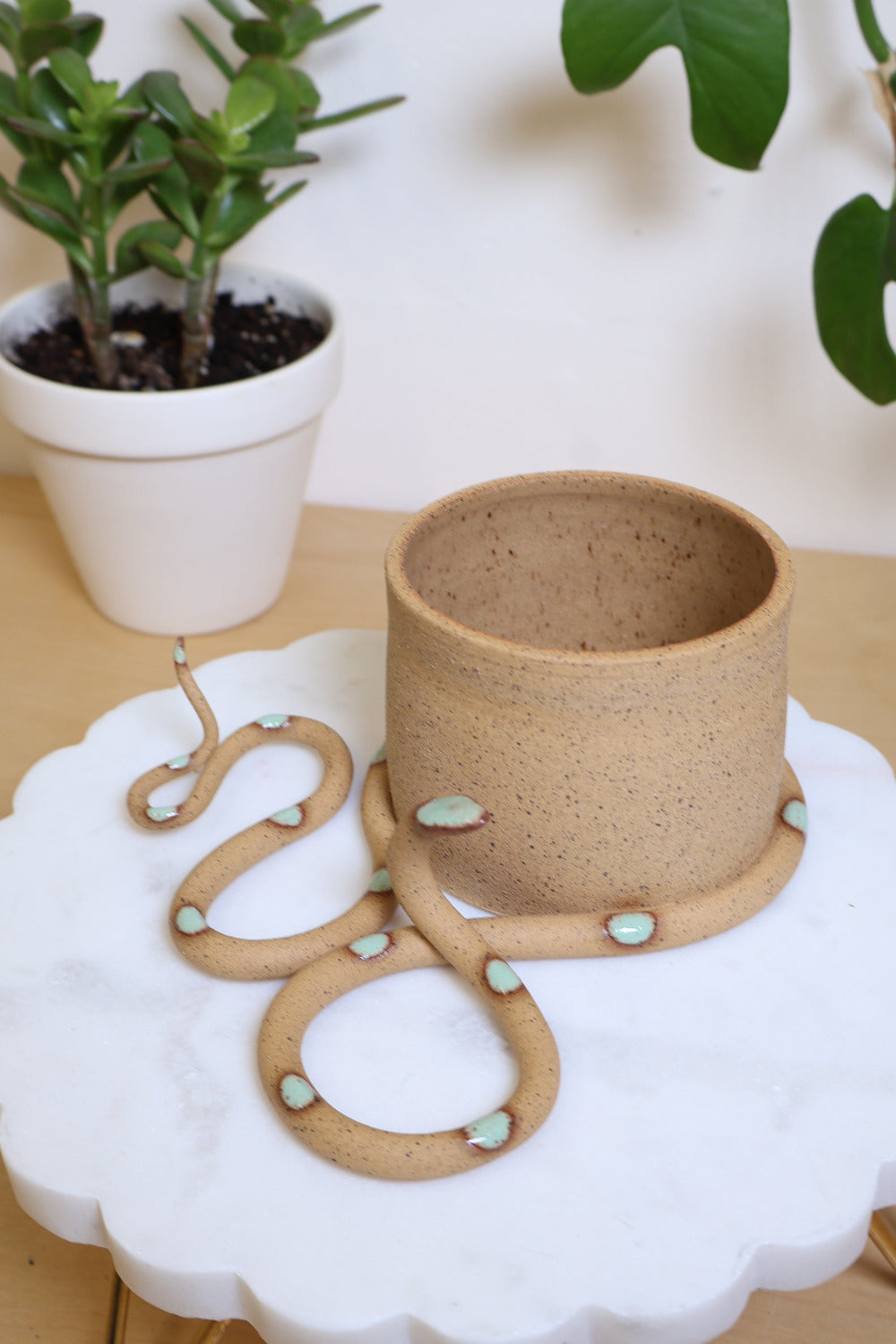 Wavy ceramic snake wrapped around a small plant pot. The snake and pot are made out of a speckled tan clay and the snake has a glazed light green head and dots. This snake is Selma. 