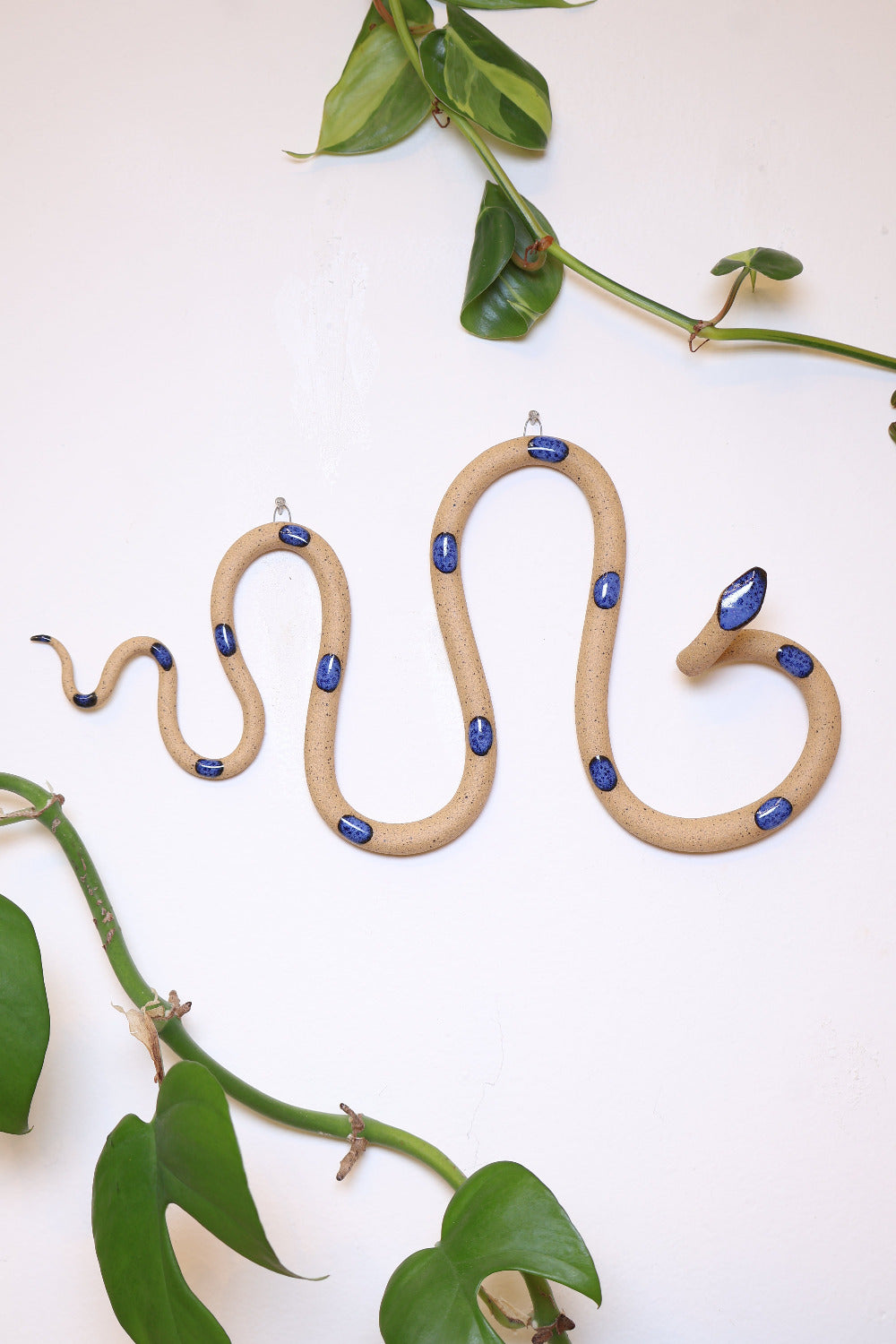 Horizontal wavy ceramic wall snake in the Selena colorway, ivory clay base and dark blue dots. It's hanging on a white wall with two small nails by its two small metal hooks.