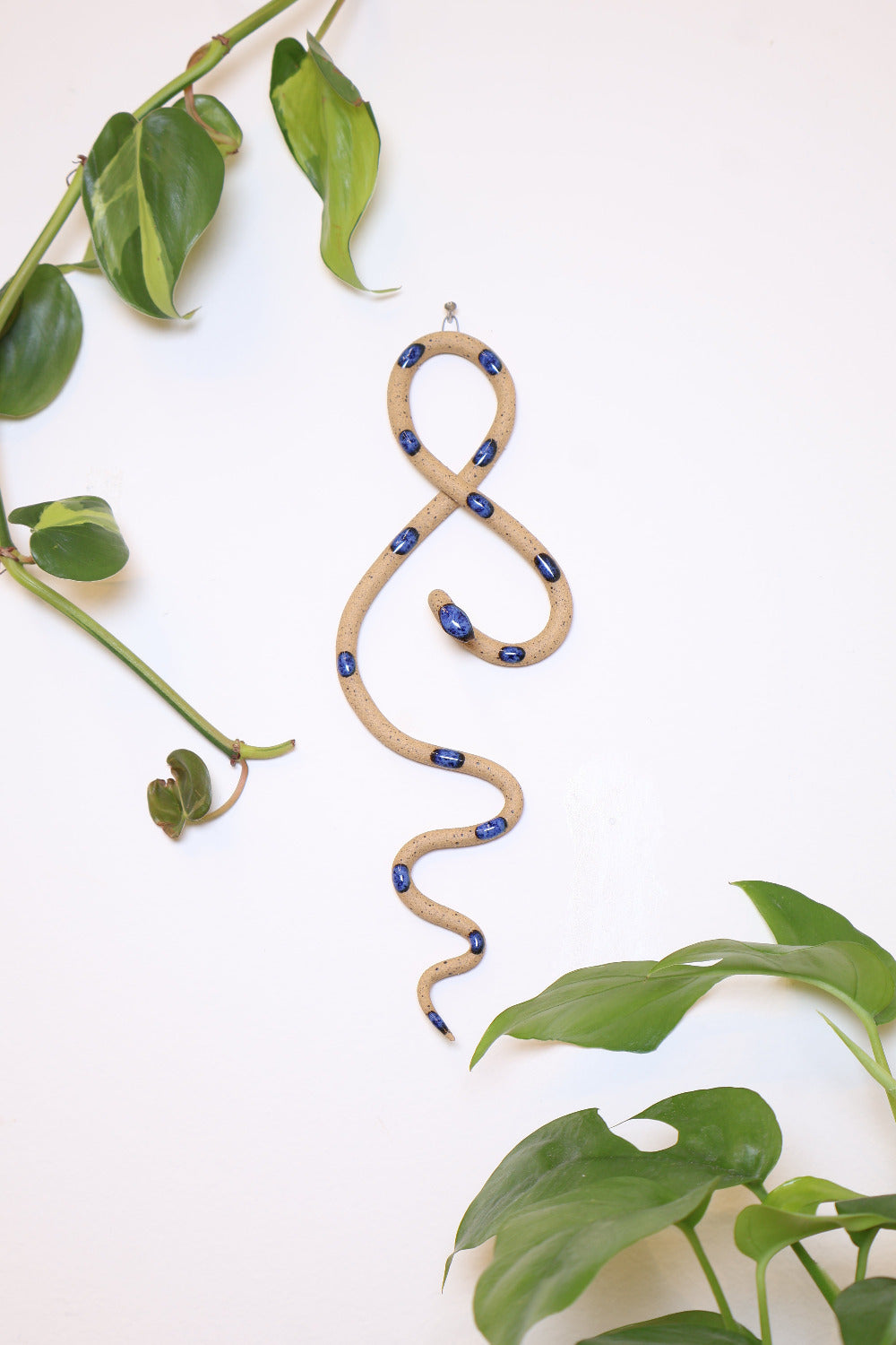Small clay snake wall sculpture/decor hanging vertically from a metal loop on a white wall. The top of the snake crosses like a figure 8 and the tail hangs down in an organic s curve. This colorway is Selena, speckled tan clay with dark blue dots. 