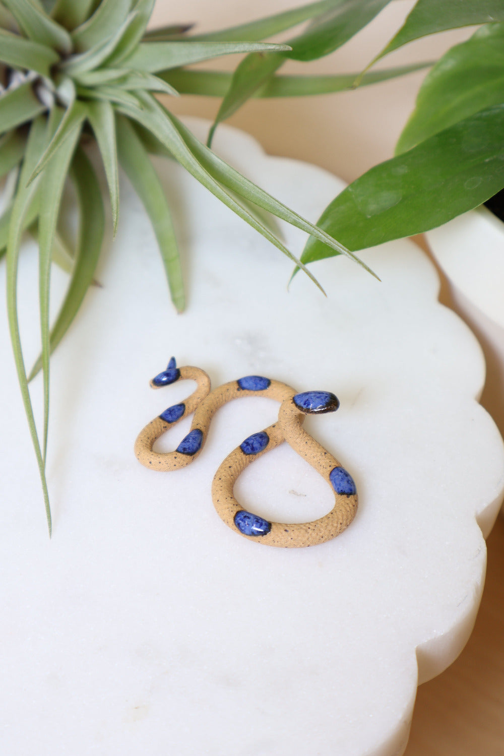 Itty bitty organic s-shaped pottery snake sculpture in the colorway Selena, speckled brown clay body with a blue glazed head and evenly spaced glazed dots along its body.