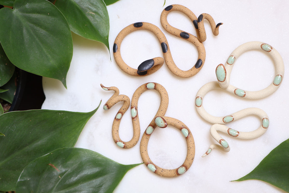 Three decorative ceramic snake sculptures on a white table surrounded by green leaves. The snake sculptures are tan or ivory clay and have green or black glazed dots. 