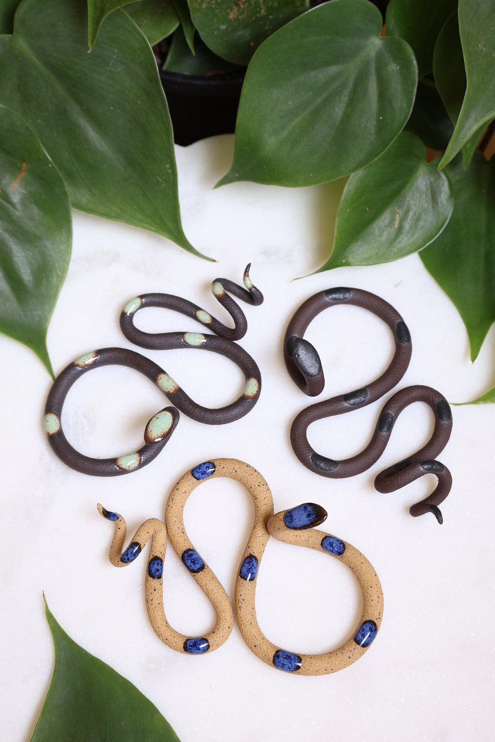 Forest curated set of three small ceramic snakes in the following colorways, Stevie (black clay with light turquoise glaze dots), Saul (black clay with black glaze dots), and Selena (speckled tan with blue glaze dots).