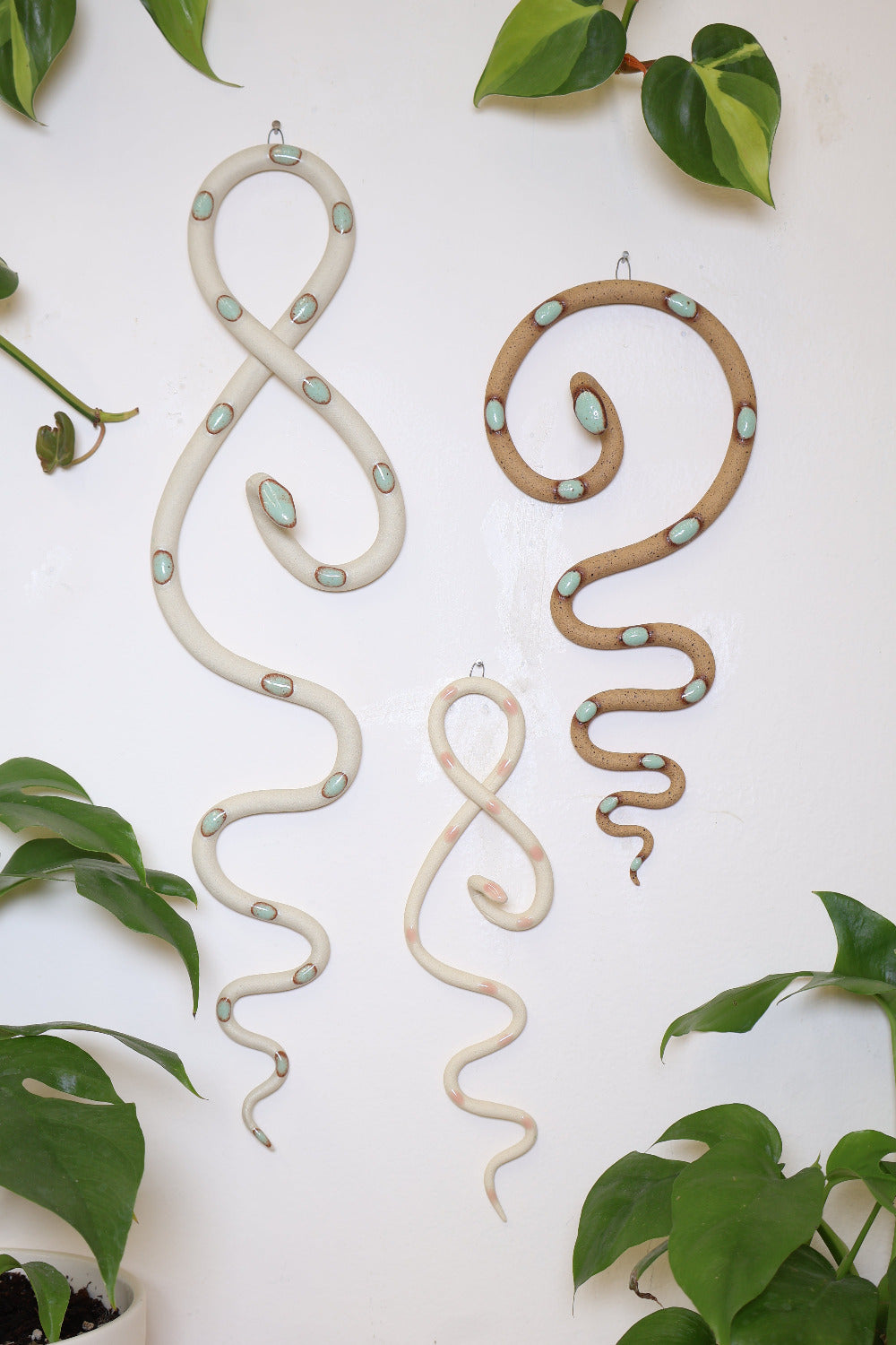 Garden curated set of three wall ceramic snakes. From left to right hanging on a wall is a  Sylvia wall snake (white clay, turquoise dots, a Sofie mini wall snake (ivory clay, pink dots) and a Selma medium wall snake (tan clay, turquoise dots).