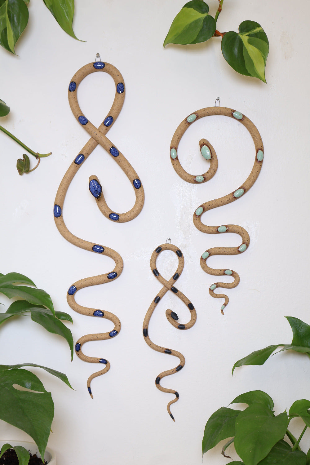 Shore curated set of three wall ceramic snakes. From left to right hanging on a wall is a Selena wall snake (speckled tan clay and blue glaze), a mini Sammy wall snake (speckled tan and black glaze), and a medium Selma wall snake (speckled tan clay and turquoise glaze).